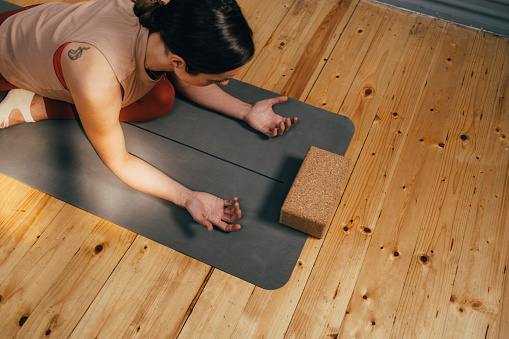 Woman leaning forward in a seated stretch on a gray yoga mat beside a cork block on a light wooden floor